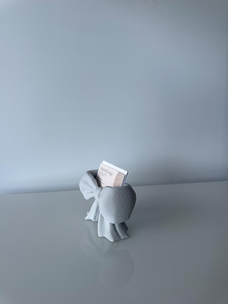 Puede incluir: Un tarjetero de color gris claro con forma de lazo, hecho de un material mate y texturizado. El soporte cuenta con una ranura central que contiene una tarjeta de visita blanca con el texto Mishell Design impreso. El dise&ntilde;o imita un lazo.