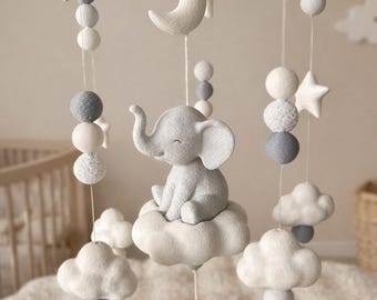 May include: A baby mobile featuring a gray elephant sitting on a white cloud, with hanging stars, moons, and spheres. The mobile is suspended from a wooden ring with a neutral color palette, perfect for a nursery.