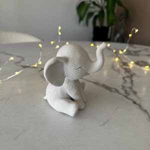 May include: A small, white elephant figurine in a seated position. The elephant has large ears, a curled trunk, and a smiling expression. The figurine is on a white marble surface, with a string of small yellow lights in the background.