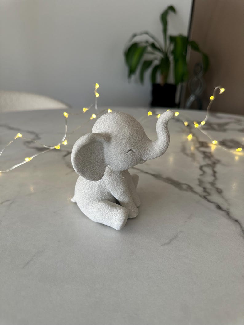 May include: A small, white elephant figurine in a seated position. The elephant has large ears, a curled trunk, and a smiling expression. The figurine is on a white marble surface, with a string of small yellow lights in the background.