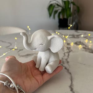 May include: A small, white elephant figurine with a textured surface. The elephant is in a seated position with its trunk curled and eyes closed. The figurine is held in a hand, suggesting its small size. The overall theme is a decorative item.