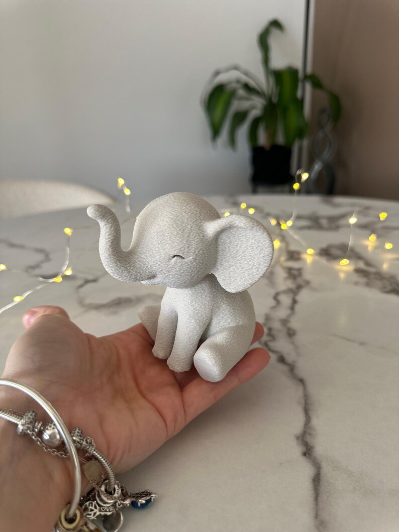 May include: A small, white elephant figurine with a textured surface. The elephant is in a seated position with its trunk curled and eyes closed. The figurine is held in a hand, suggesting its small size. The overall theme is a decorative item.