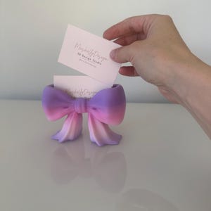 Puede incluir: Un tarjetero con forma de lazo que presenta un degradado de color desde el morado en la parte superior hasta el rosa y melocot&oacute;n claro en la base. El soporte contiene una tarjeta de visita blanca con el texto MushellyDesign 3D Design Studio.