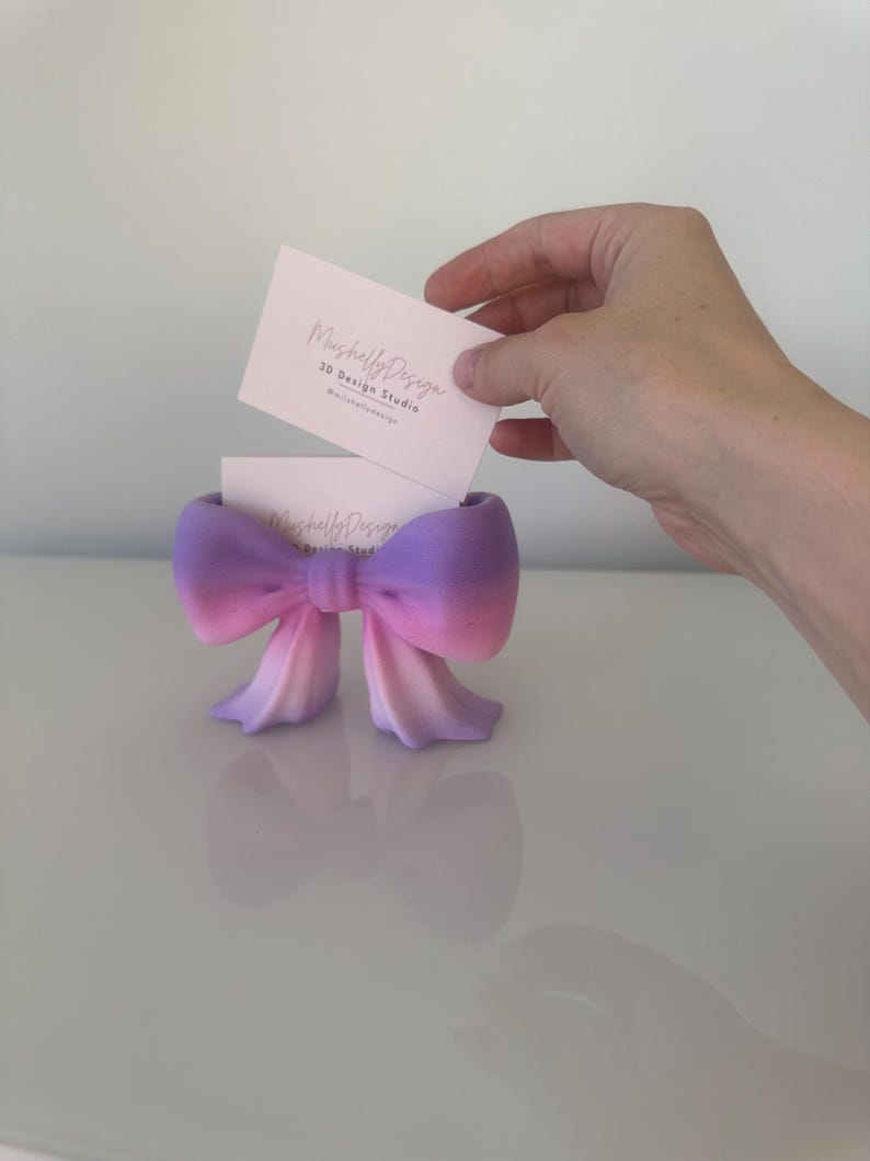 Puede incluir: Un tarjetero con forma de lazo que presenta un degradado de color desde el morado en la parte superior hasta el rosa y melocot&oacute;n claro en la base. El soporte contiene una tarjeta de visita blanca con el texto MushellyDesign 3D Design Studio.
