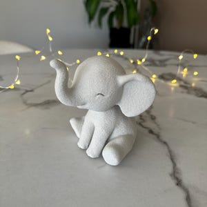 May include: A small, white elephant figurine with a textured surface. The elephant is sitting with its trunk curled up and its eyes closed. The figurine is on a marble-like surface, with string lights in the background.