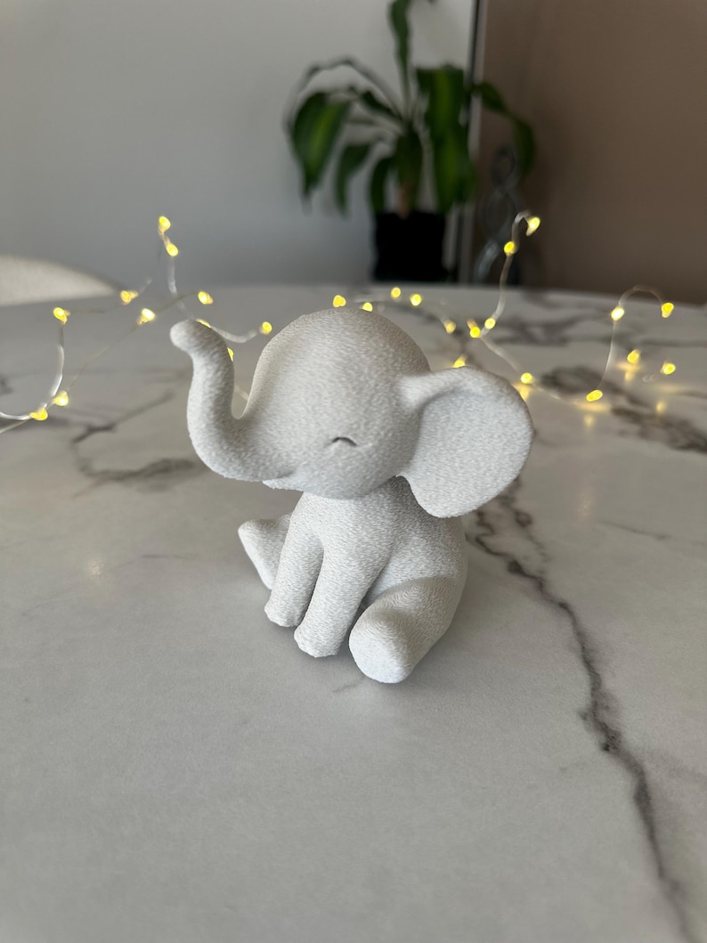 May include: A small, white elephant figurine with a textured surface. The elephant is sitting with its trunk curled up and its eyes closed. The figurine is on a marble-like surface, with string lights in the background.