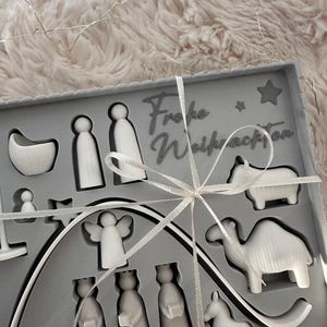 Puede incluir: Un juego de caja gris que contiene figuras blancas del bel&eacute;n, incluyendo figuras, animales y estrellas. La caja est&aacute; atada con una cinta blanca y presenta el texto "Frohe Weihnachten".