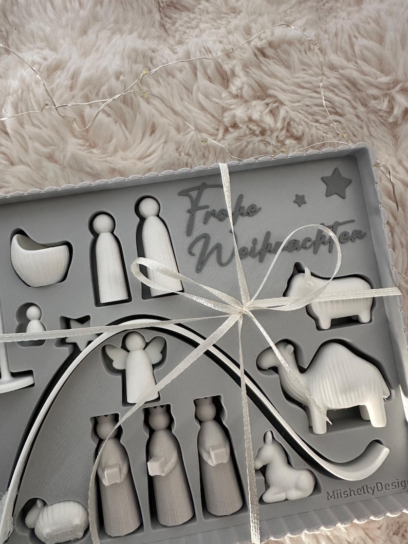Puede incluir: Un juego de caja gris que contiene figuras blancas del bel&eacute;n, incluyendo figuras, animales y estrellas. La caja est&aacute; atada con una cinta blanca y presenta el texto "Frohe Weihnachten".