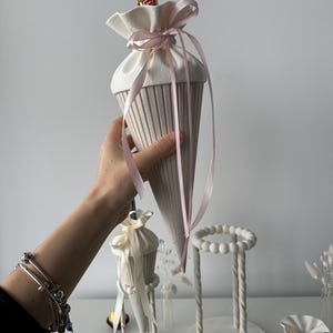 K&ouml;nnte beinhalten: Eine cremefarbene, kegelf&ouml;rmige Geschenkt&uuml;te mit gerippter Textur, die oben mit einem hellrosa Band verschlossen ist. Die T&uuml;te enth&auml;lt einen Schokoriegel. Daneben stehen wei&szlig;e St&auml;nder mit der Aufschrift 'SCHULKIND' und '2026'.