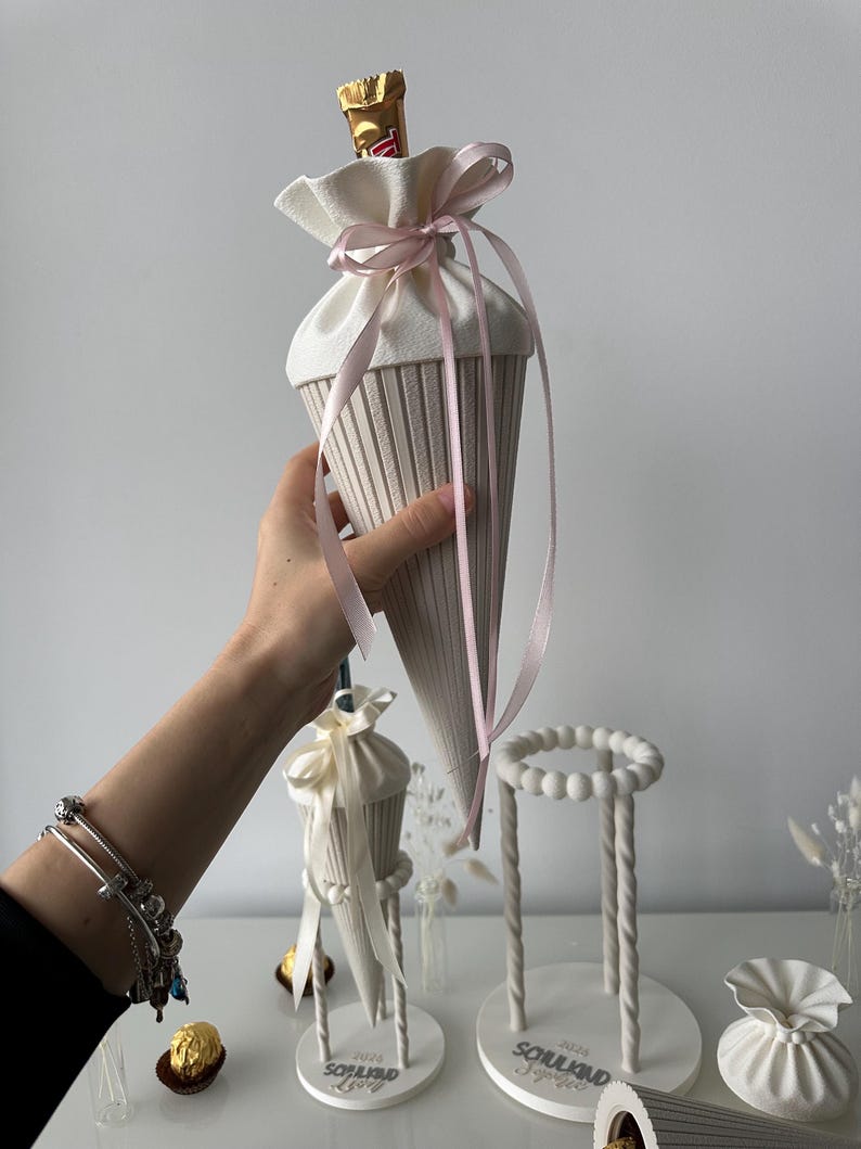 K&ouml;nnte beinhalten: Eine cremefarbene, kegelf&ouml;rmige Geschenkt&uuml;te mit gerippter Textur, die oben mit einem hellrosa Band verschlossen ist. Die T&uuml;te enth&auml;lt einen Schokoriegel. Daneben stehen wei&szlig;e St&auml;nder mit der Aufschrift 'SCHULKIND' und '2026'.