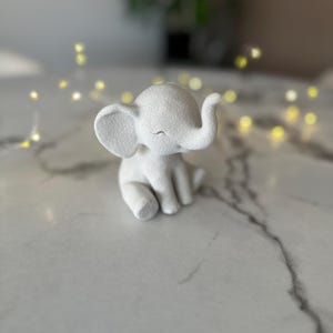 May include: A small, white elephant figurine with a textured surface. The elephant is sitting with its eyes closed and trunk raised. The figurine is on a white marble surface, with blurred yellow lights in the background.