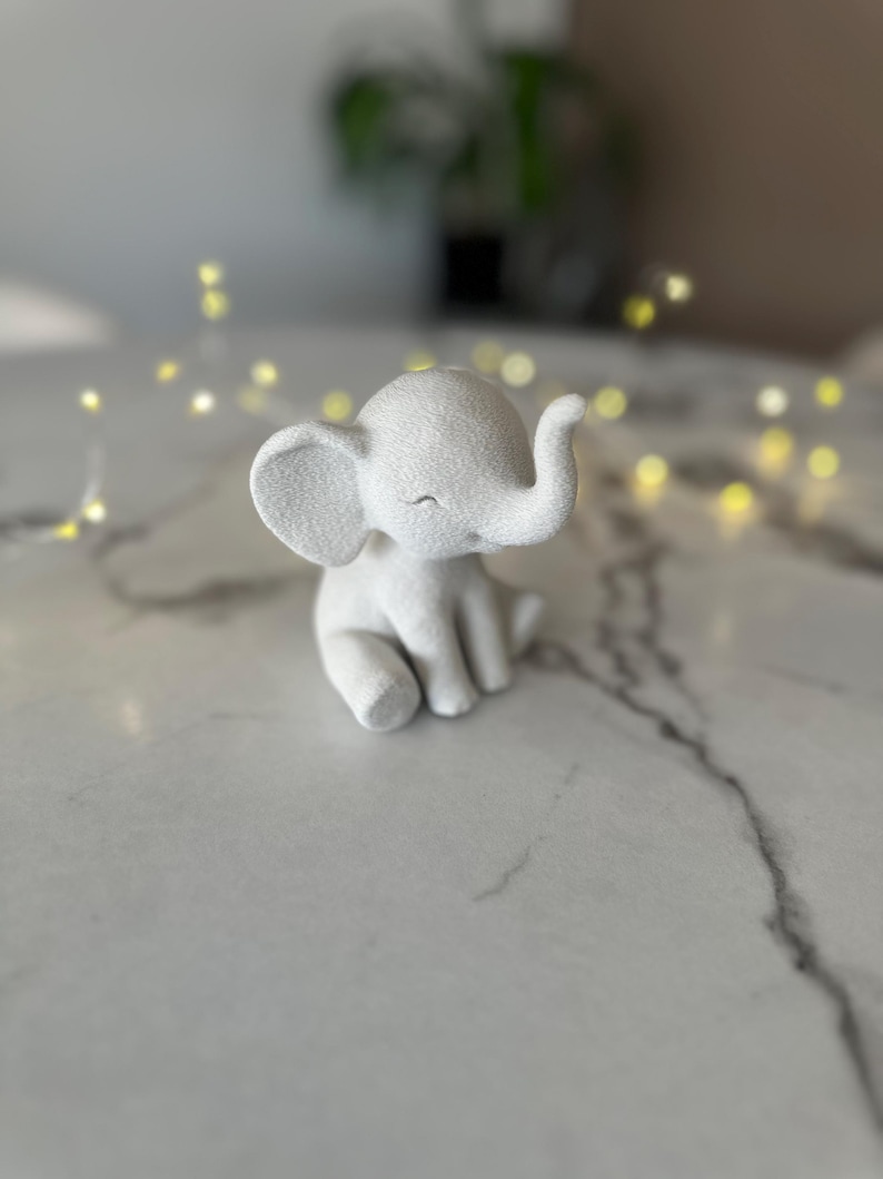 May include: A small, white elephant figurine with a textured surface. The elephant is sitting with its eyes closed and trunk raised. The figurine is on a white marble surface, with blurred yellow lights in the background.