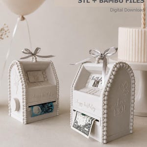 Puede incluir: Dos dispensadores de dinero en forma de cajero automático, impresos en 3D en color blanco roto, con bordes de cuentas, una cinta plateada superior y una perilla lateral. El frente muestra el texto 'happy birthday' y 'make a wish' con una ranura.