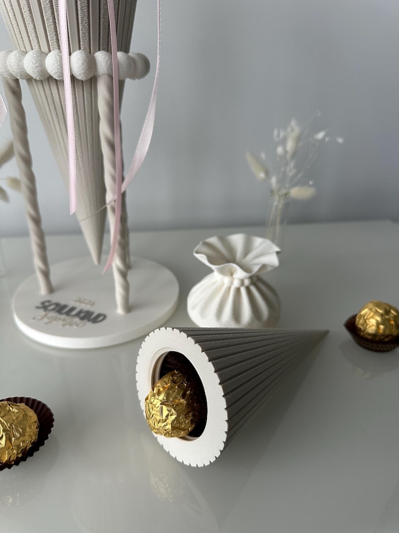 K&ouml;nnte beinhalten: Ein hellgrauer, gerippter, kegelf&ouml;rmiger Beh&auml;lter mit einer goldverpackten Schokoladentr&uuml;ffel. Ein gr&ouml;&szlig;erer, passender kegelf&ouml;rmiger St&auml;nder mit rosa B&auml;ndern und dem Text "2025" steht daneben, zusammen mit einem kleinen wei&szlig;en Beutel und weiteren Goldtr&uuml;ffeln.