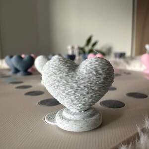 May include: A gray, heart-shaped decorative object with a textured surface, resting on a round base. The heart is the focal point, with other heart-shaped objects in the background. The object is on a light-colored surface.