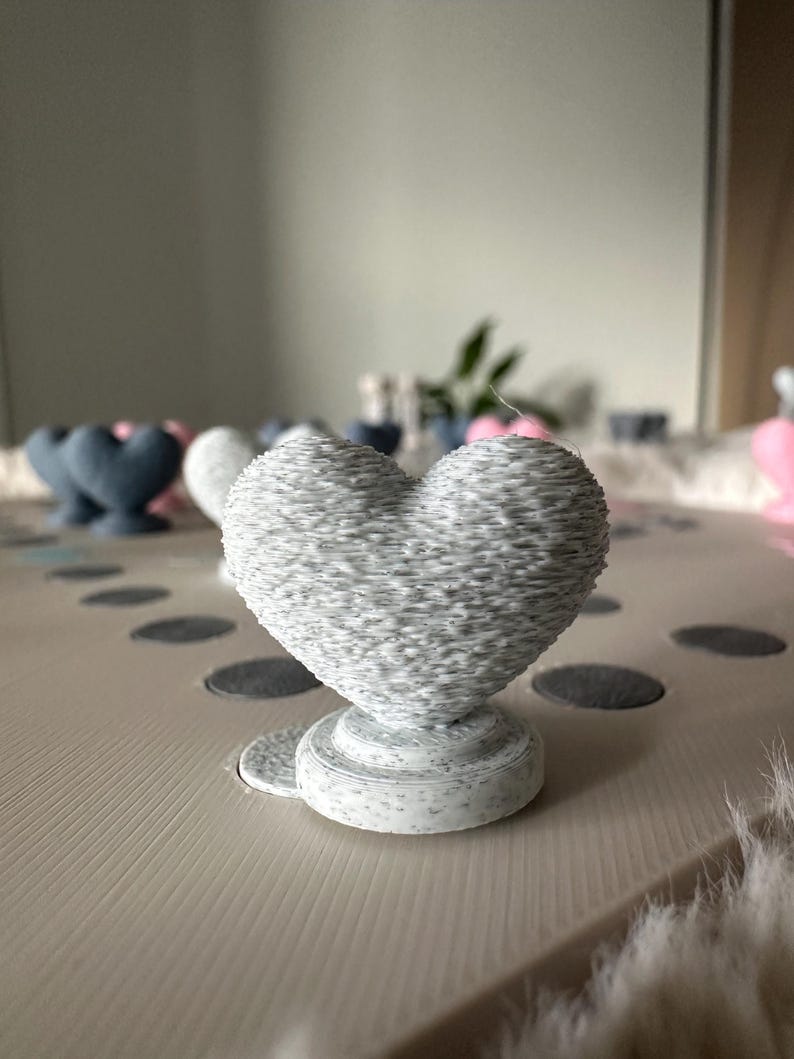 May include: A gray, heart-shaped decorative object with a textured surface, resting on a round base. The heart is the focal point, with other heart-shaped objects in the background. The object is on a light-colored surface.