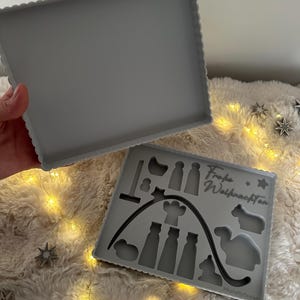 Puede incluir: Dos moldes de silicona grises, uno rectangular y otro con formas navide&ntilde;as. El segundo molde presenta siluetas de figuras, estrellas y el texto "Frohe Weihnachten". Los moldes tienen un borde festoneado.