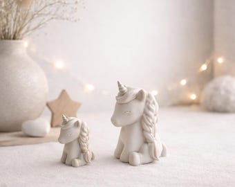 May include: Two white ceramic unicorn figurines, one large and one small, sit side-by-side. The unicorns have braided manes and small horns. The background includes a vase with dried flowers, a wooden star, and soft lighting.