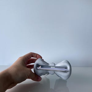 Puede incluir: Un tarjetero en forma de lazo de color gris claro con una superficie texturizada. El objeto cuenta con una ranura horizontal central dise&ntilde;ada para sujetar de forma segura art&iacute;culos de papel finos como tarjetas de visita o notas.