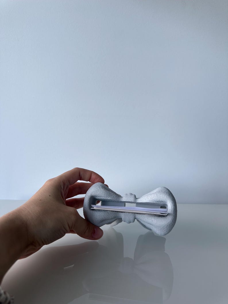 Puede incluir: Un tarjetero en forma de lazo de color gris claro con una superficie texturizada. El objeto cuenta con una ranura horizontal central dise&ntilde;ada para sujetar de forma segura art&iacute;culos de papel finos como tarjetas de visita o notas.