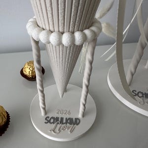 K&ouml;nnte beinhalten: Ein beigefarbener, kegelf&ouml;rmiger Geschenkkonus mit gerippter Textur, verziert mit einem Ring aus wei&szlig;en Pompons und zwei gedrehten St&uuml;tzen. Der runde Sockel tr&auml;gt die erhabene Aufschrift 2026 SCHULKIND Lion, erg&auml;nzt durch zwei goldene Pralinen.