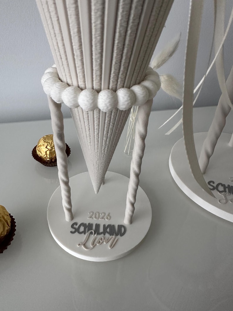 K&ouml;nnte beinhalten: Ein beigefarbener, kegelf&ouml;rmiger Geschenkkonus mit gerippter Textur, verziert mit einem Ring aus wei&szlig;en Pompons und zwei gedrehten St&uuml;tzen. Der runde Sockel tr&auml;gt die erhabene Aufschrift 2026 SCHULKIND Lion, erg&auml;nzt durch zwei goldene Pralinen.