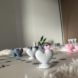 May include: A collection of heart-shaped decorations in various colors, including gray, pink, and blue. The hearts are displayed on a light-colored surface with scattered confetti. Some hearts are on pedestals.