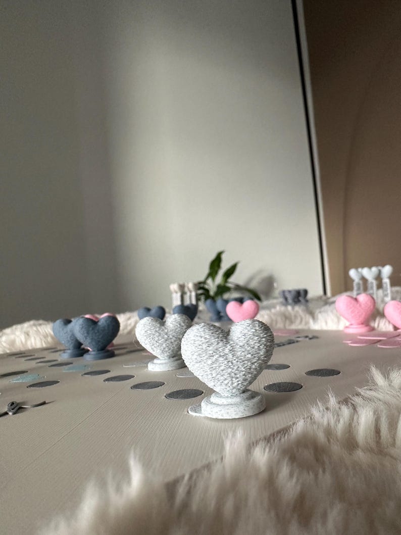 May include: A collection of heart-shaped decorations in various colors, including gray, pink, and blue. The hearts are displayed on a light-colored surface with scattered confetti. Some hearts are on pedestals.