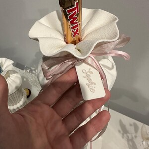 K&ouml;nnte beinhalten: Ein wei&szlig;er Stoffbeutel mit ger&uuml;schter &Ouml;ffnung, der einen Twix-Schokoriegel enth&auml;lt. Der Beutel ist mit einem hellrosa Satinband zu einer Schleife gebunden und mit einem wei&szlig;en rechteckigen Anh&auml;nger versehen, auf dem der Name Sophie eingepr&auml;gt ist.