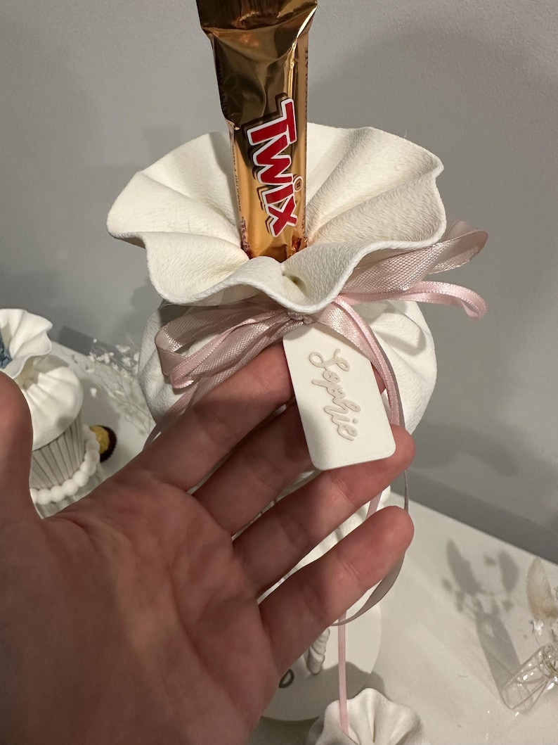 K&ouml;nnte beinhalten: Ein wei&szlig;er Stoffbeutel mit ger&uuml;schter &Ouml;ffnung, der einen Twix-Schokoriegel enth&auml;lt. Der Beutel ist mit einem hellrosa Satinband zu einer Schleife gebunden und mit einem wei&szlig;en rechteckigen Anh&auml;nger versehen, auf dem der Name Sophie eingepr&auml;gt ist.