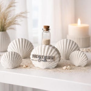 May include: A collection of four white, textured, fan-shaped shell sculptures. The central shell features a small glass bottle with a cork stopper filled with sand, and the text NORDSEE liebe in raised, dark grey, three-dimensional lettering.