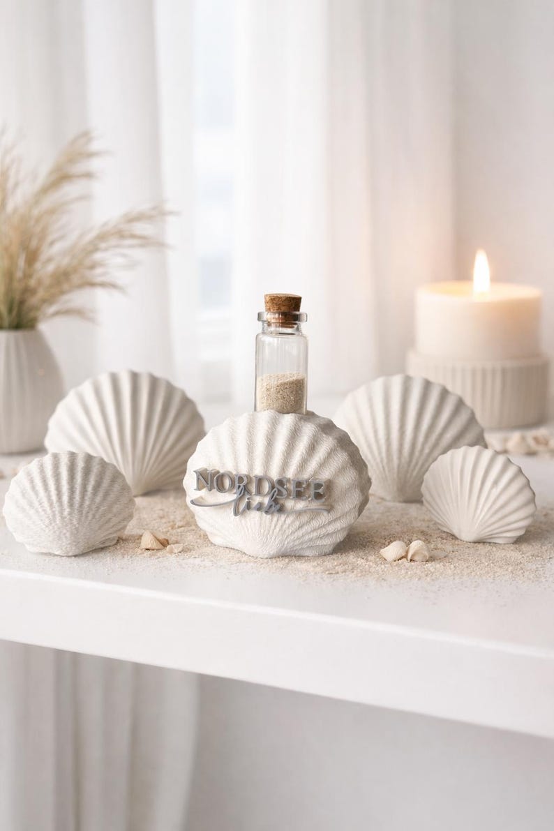 May include: A collection of four white, textured, fan-shaped shell sculptures. The central shell features a small glass bottle with a cork stopper filled with sand, and the text NORDSEE liebe in raised, dark grey, three-dimensional lettering.