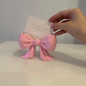 Puede incluir: Un tarjetero en forma de lazo de color rosa claro hecho de un material mate. El soporte presenta un nudo central y dos extremos de cinta colgantes. Una tarjeta de visita blanca con el texto MushellyDesign 3D Design Studio est&aacute; insertada.