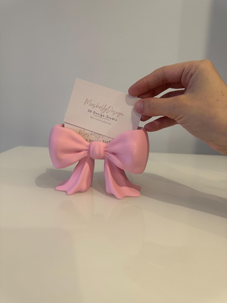Puede incluir: Un tarjetero en forma de lazo de color rosa claro hecho de un material mate. El soporte presenta un nudo central y dos extremos de cinta colgantes. Una tarjeta de visita blanca con el texto MushellyDesign 3D Design Studio est&aacute; insertada.