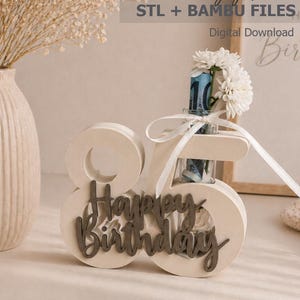 Puede incluir: Un expositor con el número 85 impreso en 3D de color crema con el texto Happy Birthday en letra cursiva marrón oscuro. Un tubo de ensayo de cristal transparente se inserta en el número 5, conteniendo un billete enrollado, flores y un lazo.