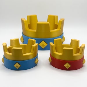 Könnte beinhalten: Drei dekorative kronenförmige Objekte in verschiedenen Größen. Die Kronen sind goldfarben mit blauen oder roten Bändern um die Basis, die jeweils mit goldfarbenen rautenförmigen Akzenten verziert sind. Der Hintergrund ist weiß.