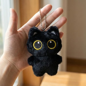 Może przedstawiać: Mały, czarny pluszowy kot z dużymi żółtymi oczami i srebrnym łańcuszkiem. Kot ma puszystą fakturę i uroczy, kreskówkowy design. Brelok jest trzymany w dłoni.