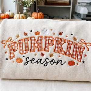 Könnte beinhalten: Ein cremefarbenes Sweatshirt mit einer Stickerei, die PUMPKIN season liest. Das Wort PUMPKIN ist in einem braun-weißen Vichy-Muster gehalten, während season in schwarzer Schreibschrift steht. Das Design enthält orange Kürbisse, Blätter, Sterne und ein Gespenst.