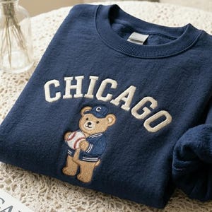 May include: Navy blue sweatshirt with the word "CHICAGO" embroidered in cream-colored thread. A cartoon bear wearing a baseball uniform and holding a baseball is embroidered below the word. The sweatshirt is folded on a table.