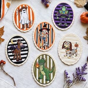 May include: Six oval-shaped embroidered patches featuring cartoon characters in Halloween costumes. The designs include a ghost, a space ranger, a cowgirl, a horse with bat wings, a mummy dog, and a dinosaur holding a flame, all on striped backgrounds.