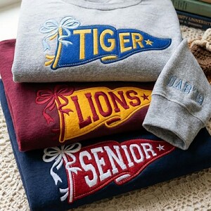 May include: Three sweatshirts in gray, burgundy, and navy blue are stacked. Each has a banner design with text: "TIGER", "LIONS", and "SENIOR". A gray cuff with "DAD '19" is next to them. A baseball, whistle, and books are in the background.