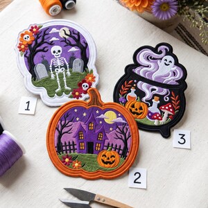 Może przedstawiać: Trzy haftowane naszywki z motywami Halloween: szkielet na cmentarzu, nawiedzony dom z dynią oraz duch unoszący się nad kociołkiem. Naszywki ułożono obok szpulek nici w kolorze pomarańczowym, czarnym i fioletowym oraz nożyczek.