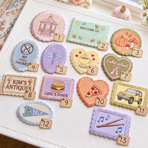 Peut inclure: Une collection de treize écussons brodés aux bords festonnés, présentant divers motifs dont un kiosque, un panneau Stars Hollow, une citrouille, un panneau interdiction de téléphones, une libellule, une tasse de café, des chaussons de danse, Kim's Antiques, un burger, une pizza, une jeep, un fanion Yale et des baguettes de batterie.