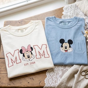 May include: An off-white t-shirt with pink embroidered text reading MOM and a Minnie Mouse graphic, and a light blue t-shirt with blue embroidered text reading DAD and a Mickey Mouse graphic. Both shirts feature the text EST. 2024 below the characters.
