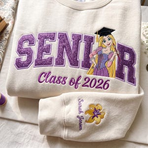 Op de afbeelding: Een crèmekleurige sweater met ronde hals en paarse glitterende geborduurde tekst met de woorden SENIOR en Class of 2026. Een geborduurd personage met lang blond haar en een afstudeerhoed is aanwezig. De mouw bevat de naam Sarah Jensen.