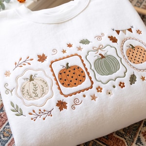 May include: A white crewneck sweatshirt featuring detailed embroidery of three pumpkins in scalloped frames. The pumpkins are orange with black hearts, sage green, and cream with vine patterns, surrounded by embroidered autumn leaves and stars.