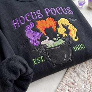Könnte beinhalten: Ein schwarzes Sweatshirt mit einer Stickerei, die den violetten Schriftzug HOCUS POCUS über drei stilisierten Haarformen in Violett, Orange und Gelb zeigt. Darunter ein schwarzer Kessel mit grünen Blasen, Spinnweben, Fledermäusen und dem Text EST. 1693.