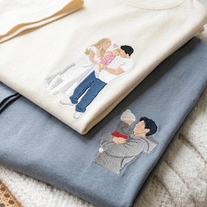 May include: Cream-colored and blue t-shirts with embroidered family portraits. One shirt features a couple holding a baby, the other a parent and child. The embroidery is detailed, with figures in various colors.