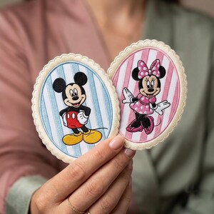 May include: Two oval-shaped embroidered patches with scalloped cream-colored edges. One patch features Mickey Mouse on a light blue striped background, while the other displays Minnie Mouse on a pink striped background, both with detailed stitching.