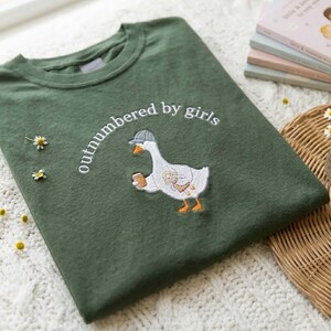 May include: Green sweatshirt with the embroidered phrase "outnumbered by girls" above a cartoon goose wearing a cap and holding a coffee cup. The goose has a rabbit design on its body. The sweatshirt is folded on a white surface.